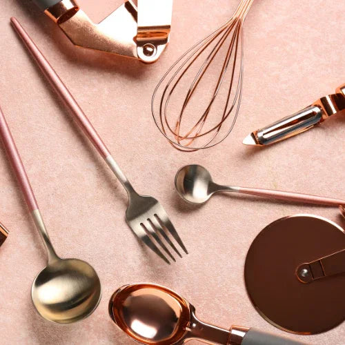 Rose gold kitchen utensils including ladle, fork, whisk, peeler, and pizza cutter on a pink background